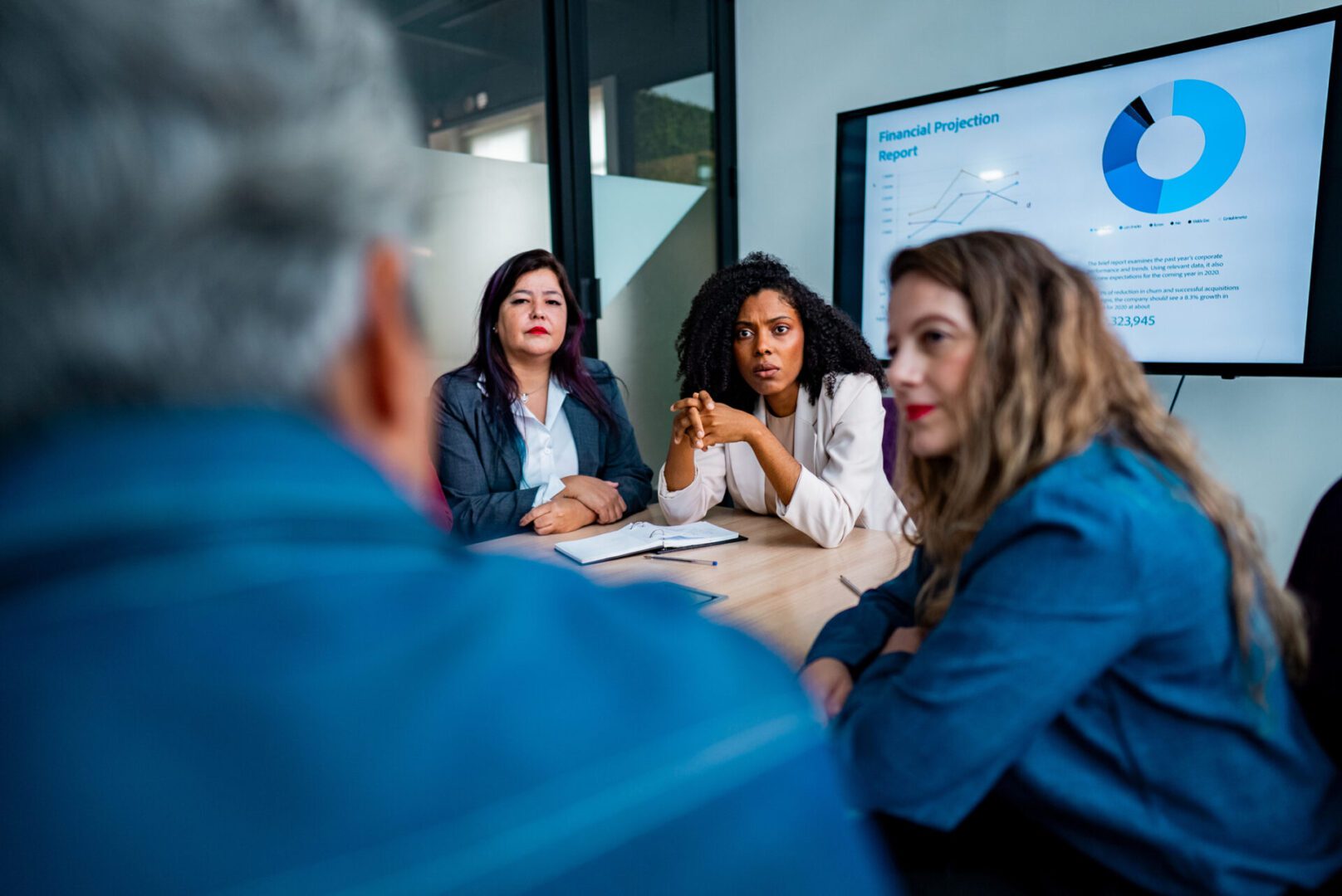 A diverse group of professionals is seated around a conference table in a modern office, engaged in a business meeting. A large screen displays charts and financial data, suggesting a strategic discussion or presentation. The setting includes laptops, notebooks, coffee mugs, and office supplies, reflecting a collaborative and analytical work environment