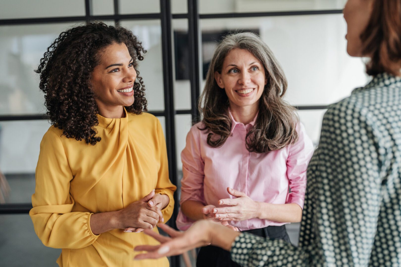 Shot of three confident business female colleagues chatting walking through the office hall, enjoy friendly warm conversation. Business women in the office having informal talk in coffee break. Smiling business persons having a conversation walking and talking